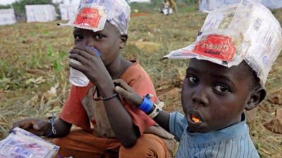 Two young refugees sit on the ground wearing plastic hats