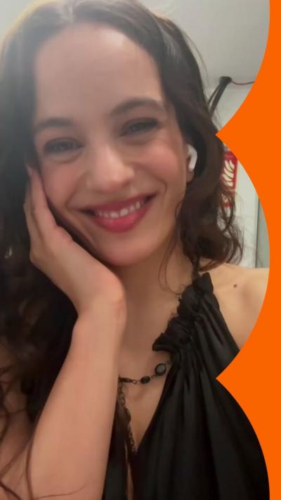 Singer Rosalia smiles with her right hand on her face. She has long brown hair and wears a black sleeveless top