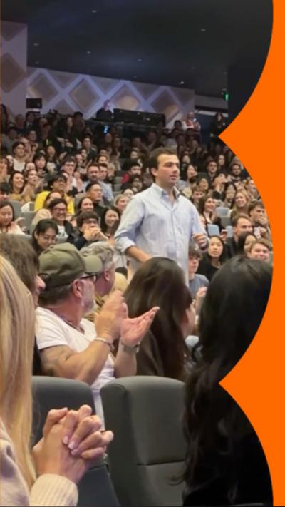 Man in a light blue shirt walking down the steps of an auditorium as the audience applause.