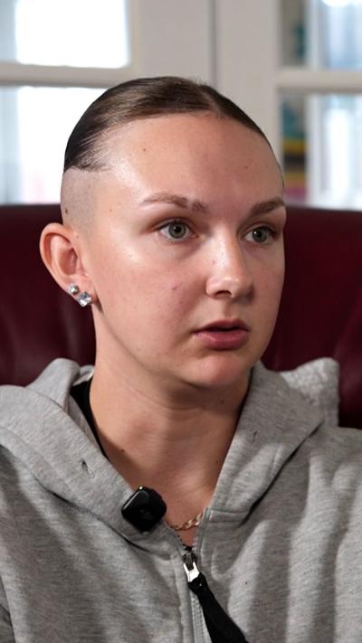 A young woman sits indoors, shown from the shoulders up. She has light skin and green eyes, with her brown hair shaved close on the sides and slicked back on top. She wears a grey zip‑up hoodie and small square stud earrings. A black microphone is clipped near her collar. The background is softly blurred, showing a maroon chair and bright daylight through a window.