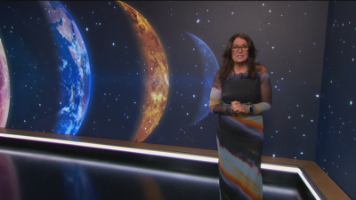 Judith Ralston in a long multi-coloured dress, standing in front of a screen with a number of planets on it