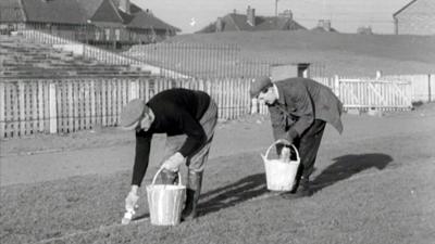 Black and white image of the groundsmen painting the lines on the pitch.  One man in flat cap and black jumper is in front, with a bucket and paintbrush, behind him another man in flat cap and jacket also with a paintbrush and bucket.  Behind there is a terrace and fence around the pitch with building visible in the background.