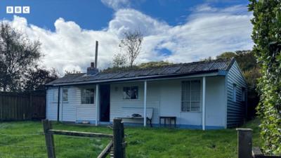 A white wooden bungalow with blue trim, it has a small front garden and is surrounded by fencing