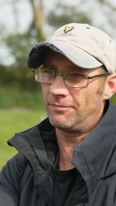 A man in a black coat and black top. He is wearing glasses and a white cap. 