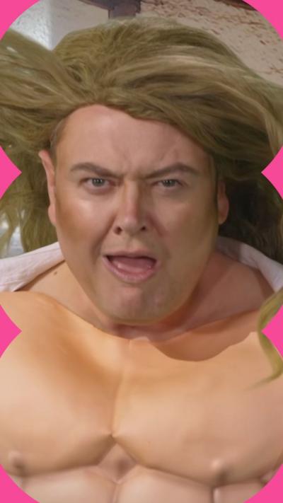 Alan Carr pulls a funny face while wearing a muscle suit and a long dark blond wig that is blowing in the wind