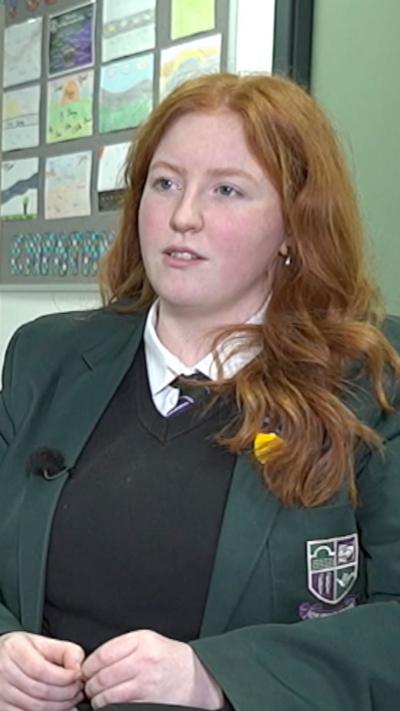 A girl with long ginger hair is smiling and looking into the camera. She is wearing a green school blazer, a tie and a white shirt. 