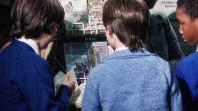 Three children in school uniform gathered around a rack of paperback books in a library.