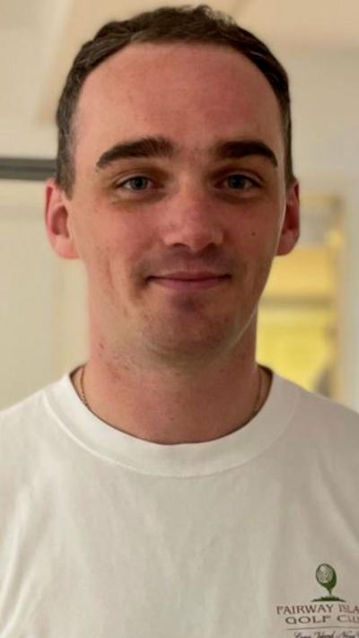 A man with short brown hair, looking at the camera. He is wearing a white t-shirt. There is a white wall and white door in the background.
