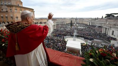 Pope Leo wears a red cape and white rope waving to crowds in Vatican City
