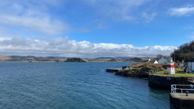 View of sea with white buildings on coastline to the right and hills in distance under a blue and cloudy sky