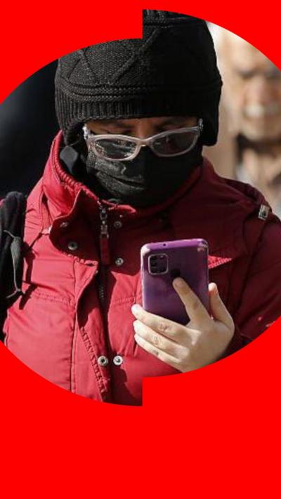 A person wear a black hat, face covering, glasses and red jacket looks at their phone