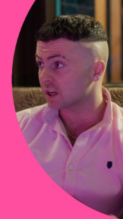 Conor has short dark hair with a skin-fade, and is wearing a pink polo-shirt.