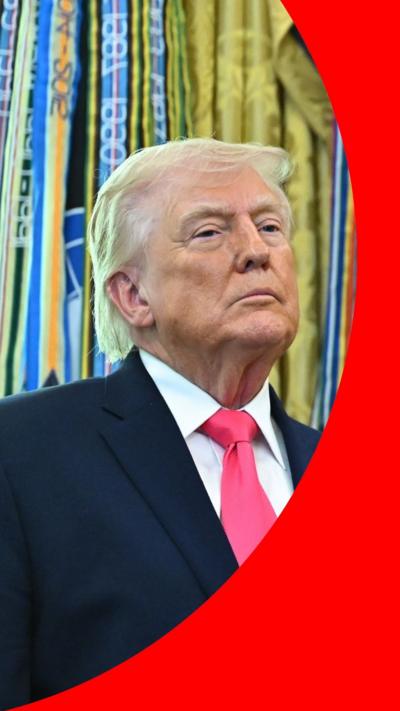 Donald Trump stands in front of brightly coloured drapes. He is a wearing a navy blue suit, white shirt and a pink tie.