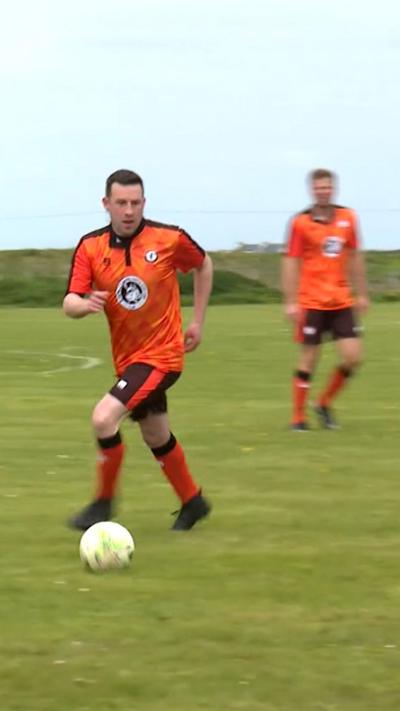 Football player dressed in orange and black running with a ball on a football pitch. There is another player in orange and black also standing behind him to the right.