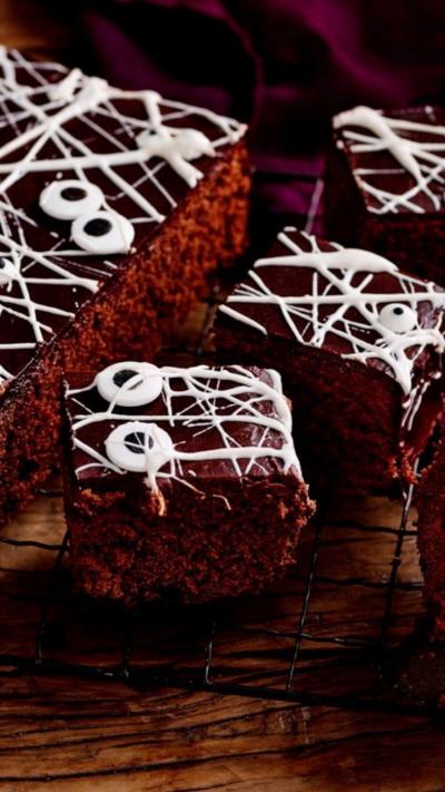 Chocolate traybake cake with white cobwebs and pairs of cartoon eyes, on a wooden table and purple cloth.