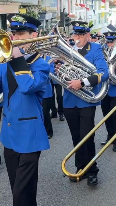 A band holding instruments. They are wearing blue suits and black trousers and hats.