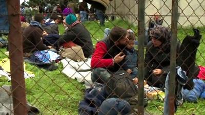 Migrants sitting on ground outside, behind fence