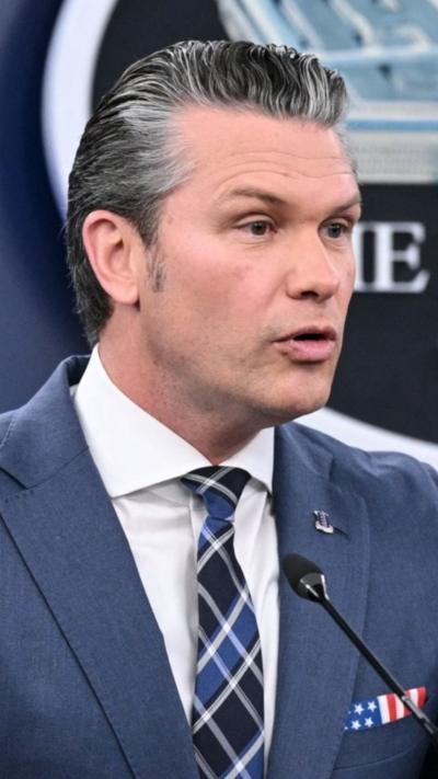 Pete Hegseth wears a light blue suit while speaking behind a podium. 