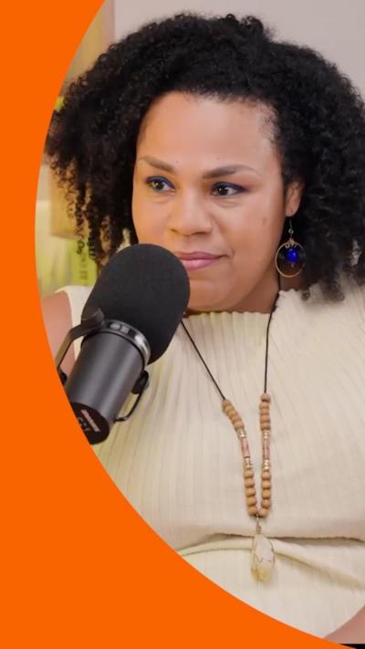 A woman with curly black hair sits behind a microphone. She is wearing a cream top and a necklace.