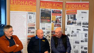 Matt Bruce, Tòrmod MacLeòid agus Alasdair MacLeòid nan suidhe a' bruidhinn agus postairean An Rudhach air an cùlaibh. 