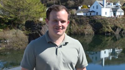 Innes MacCuidhein na sheasamh an Eilean Luinn agus gàire air aodann. 