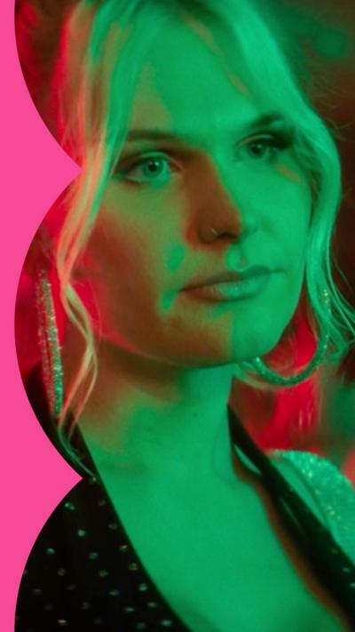 Sasha is wearing a black top. She is in a bar. Green and red lights are shining on her.