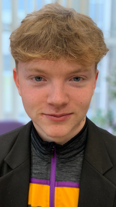 A young man with wavy fair hair looks into the camera. He has blue eyes. He is wearing a black jacket and a grey, purple and yellow scarf