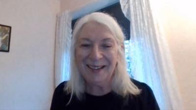 Catriona NicIlleDhuibh na suidhe air beulaibh camera agus gàire air a h-aodann. Tha falt fada glas oirre agus geansaidh dorch.