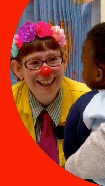 Woman in a yellow coat wears glasses, a red clown nose and flowers in her fair, looking at a young boy with his back to the camera