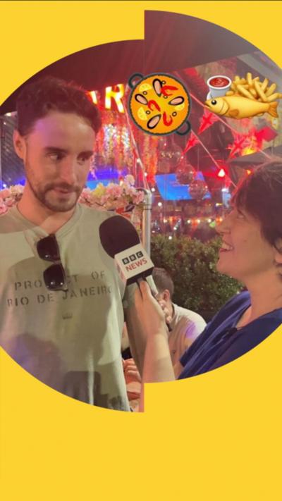 A man is interviewed by a woman in a bar. A graphic of paella and fish and chips is superimposed in the top right