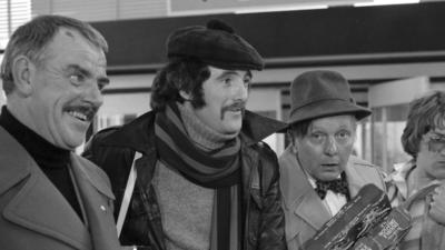 Dewis Morris pictured alongside other Grand Slam cast members. He is young, wearing a black cap and has a handlebar moustache. He is wearing a roll neck jumper, scarf and leather jacket - the whole photo is black and white. 