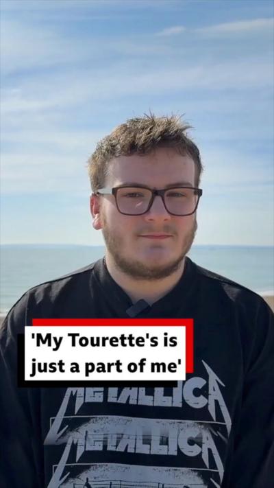 Spencer Davies-Monk on a beach wearing a black Metallica T-shirt. He is wearing black glasses and has a beard.