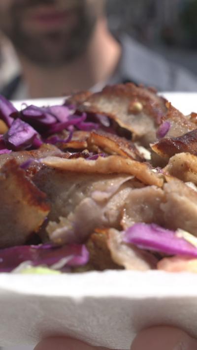 A man who is out of focus holding a kebab with brown meat, green lettuce and red (purple) cabbage. The packaging is white.
