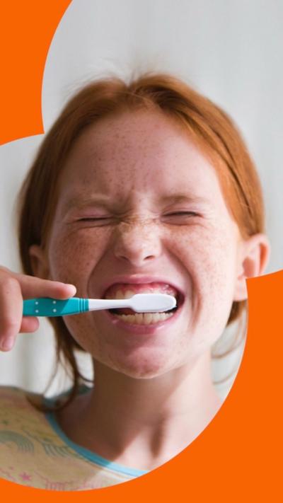Young girl brushing teeth with blue toothbrush, scrunching up her eyes, playfully.