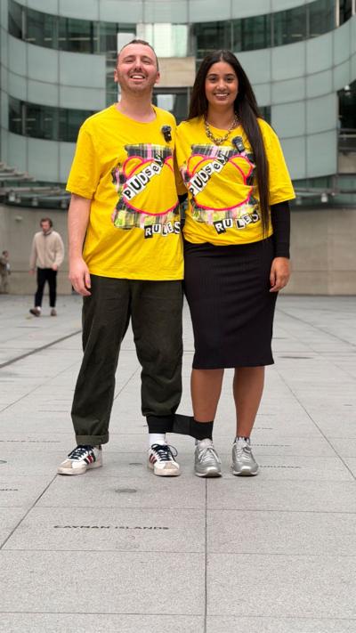 Two individuals stand together in a plaza, wearing bright yellow Children in Need branded t-shirts, surrounded by a busy urban environment. Their legs are tied together.