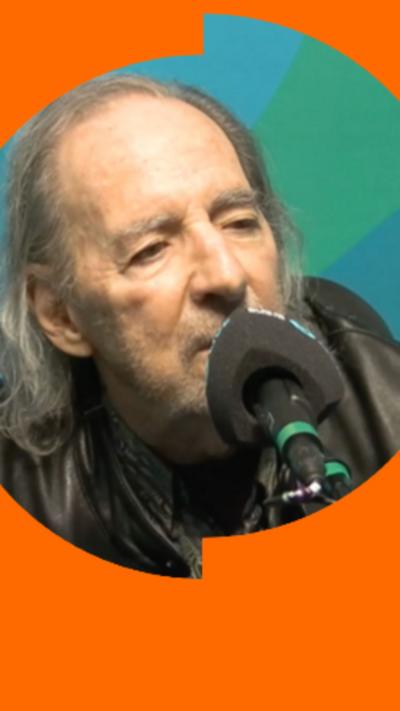 Harry Shearer has long grey hair and is wearing a black leather jacket. He is sitting in front of a microphone.