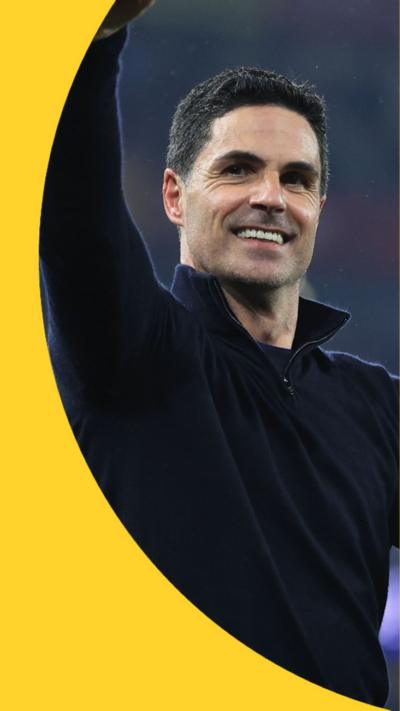 Mikel Arteta has short dark hair and is wearing a black top. He has his right arm up in the air and is smiling.