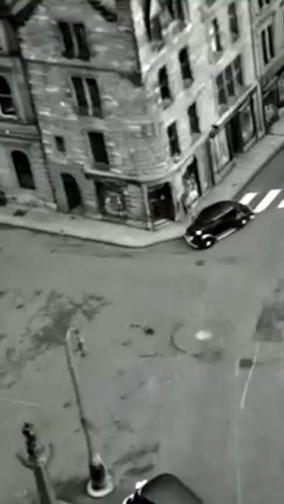 Black and white image of an old car turning a corner on an empty street in front of stone, four storey buildings.