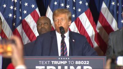 Donald Trump speaking into a microphone in front of a man and the US flag