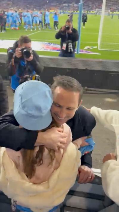 A young girl in a sky blue hat hugged by a football manager in a dark jacket