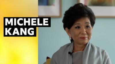 Michele Kang smiling. She is wearing a grey dress, and behind her is a grey background with the bottom halves of two photos either side of her. The caption to the left reads: "Michele Kang."