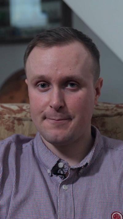 A man with short brown hair is wearing a purple shirt looking into the camera. He is sat in a large yellow/cream arm chair with red designs on it. There is a blue/white wall in the background.