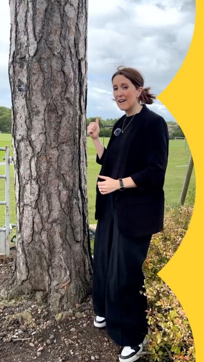 A woman stands in front of a tree and points at a field