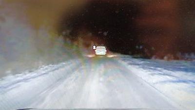 A blurry image of a snow-covered road with a white or silver van on the carriageway ahead. The sky is dark. Snow covers the verge on either side of the road.