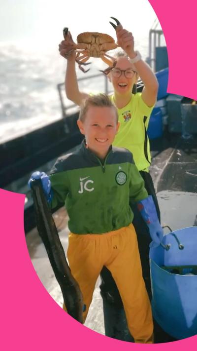 A girl and a boy stand on a boat - she is holding up a crab above his head