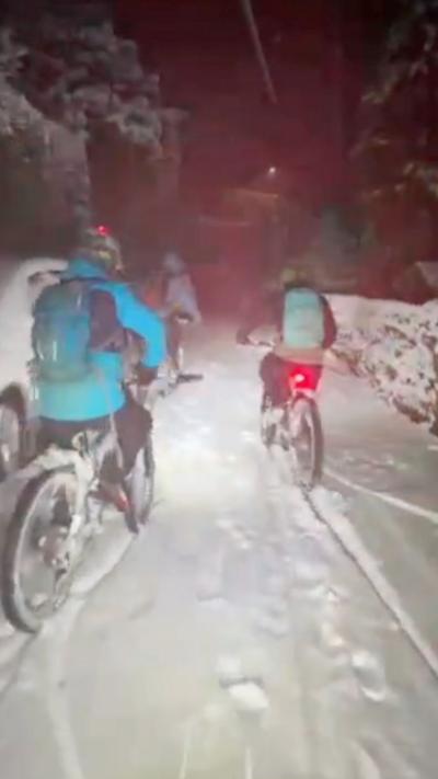 Cyclists bike down a narrow snow covered street in whilst snow falls