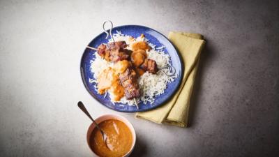 Pork skewers and rice with satay sauce on top on a blue plate. A bowl of satay sauce sits alongside.
