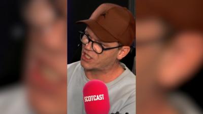 Man with a brown cap and black glasses is in front of a black background and speaking into a red 'Scotcast' microphone.