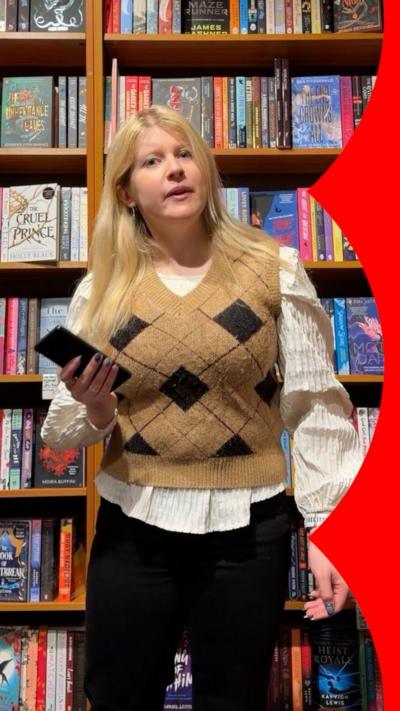 A young woman in a mustard checked top and white shirt, with blonde hair, in a book shop.