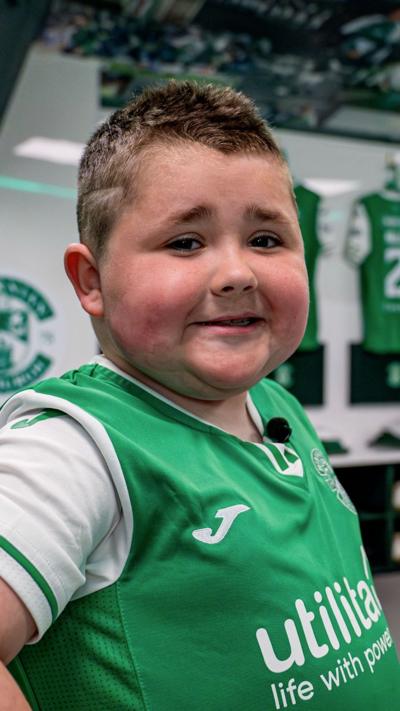 Sam Millar, eight, stands in the dressing room of Hiberian Football Club along with Dwight Gayle, one of the players. Sam is wearing a green football top and Dwight is wearing a black gilet jacket.